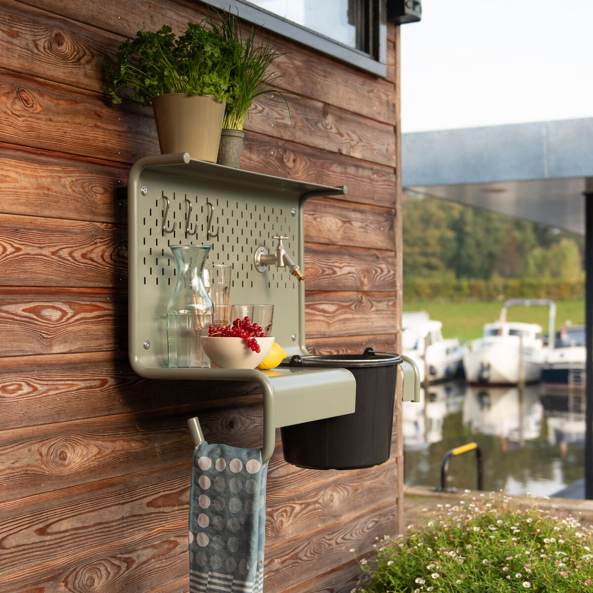 Die Gartenstation »Waterworks« von Weltevree ist an einer Holzhauswand montiert. Sie besteht aus Metall in Grün-Grau, integriertem Waschbecken, schwarzem Eimer, polierter Oberfläche, Ablage mit Kräutertopf, Wasserkaraffe, Tuchhalter und Dekoschale. Im Hintergrund ein Wasserblick.