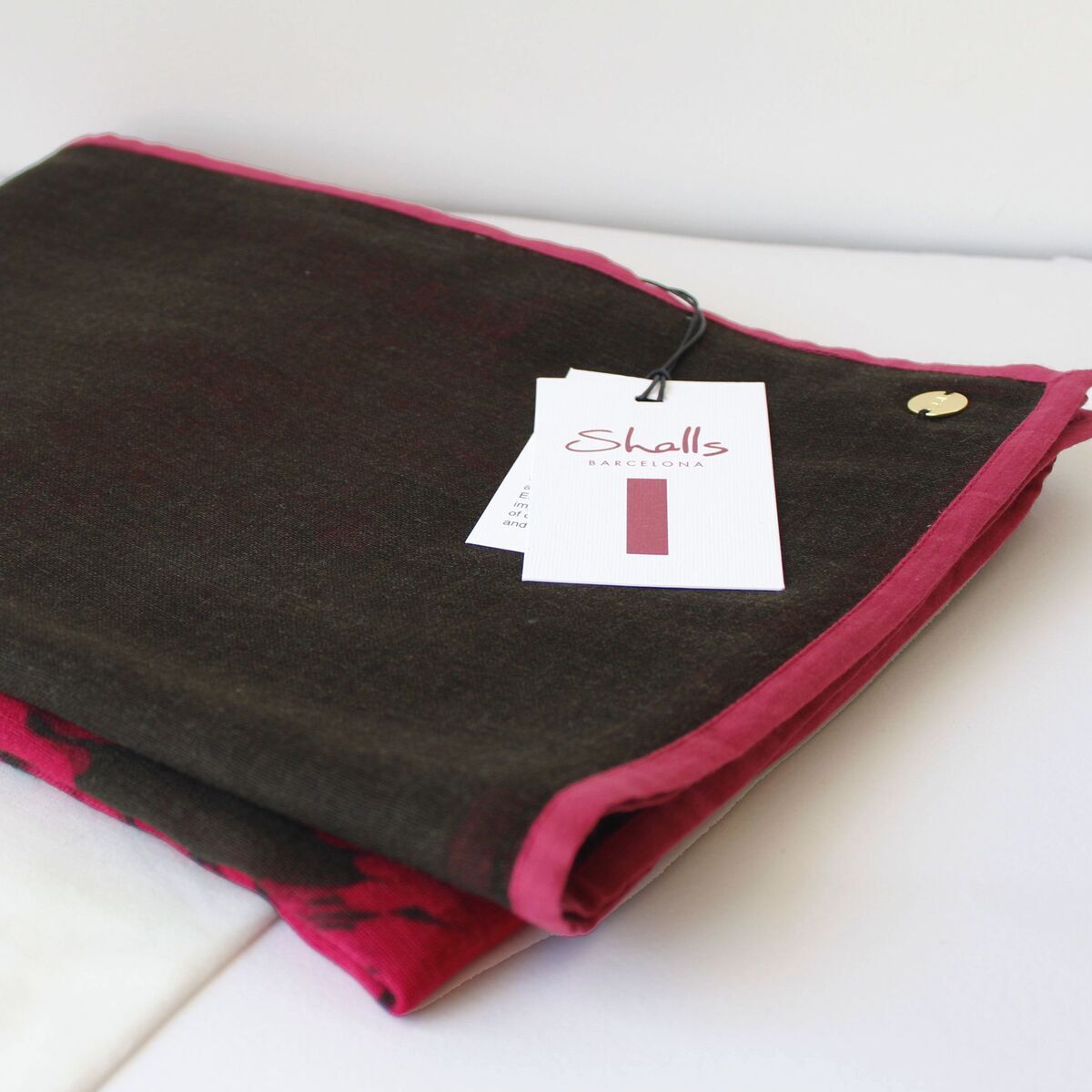 Gefalteter Wollschal in Rot- und Brauntönen, schlichter, eleganter Look mit angenähter Etikette, auf weißem Hintergrund liegend. Ein kleines, rundes, goldenes Detail ist am Schal befestigt, daneben ein weißes Label mit Produktinformation.
