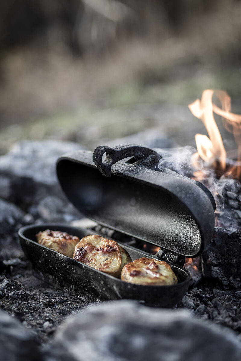 Ein Kartoffelbräter aus schwarzem Gusseisen steht auf rauchender Kohle, drei gebackene Kartoffeln sind darin zusehen. Im Hintergrund brennt ein kleines Feuer. Die Szene wirkt rustikal und einladend.