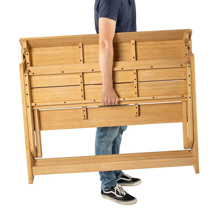 Eine Person trägt eine klappbare Tischbank aus Bambus von Weltevree. Die Sitzbank ist zusammengeklappt, zeigt stabile Holzstruktur und klare Linien. Das Möbelstück wirkt robust, handlich und eignet sich für den Innen- und Außenbereich.