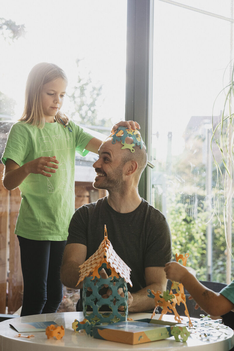 Ein Mann und zwei Kinder spielen mit dem Spiel »Binabo« (60 Teile) an einem Tisch. Ein Kind setzt dem Mann spielerisch Bauteile auf den Kopf. Das Spiel besteht aus farbigen, biegsamen Teilen, die zusammengesetzt werden können, um dreidimensionale Strukturen zu schaffen.