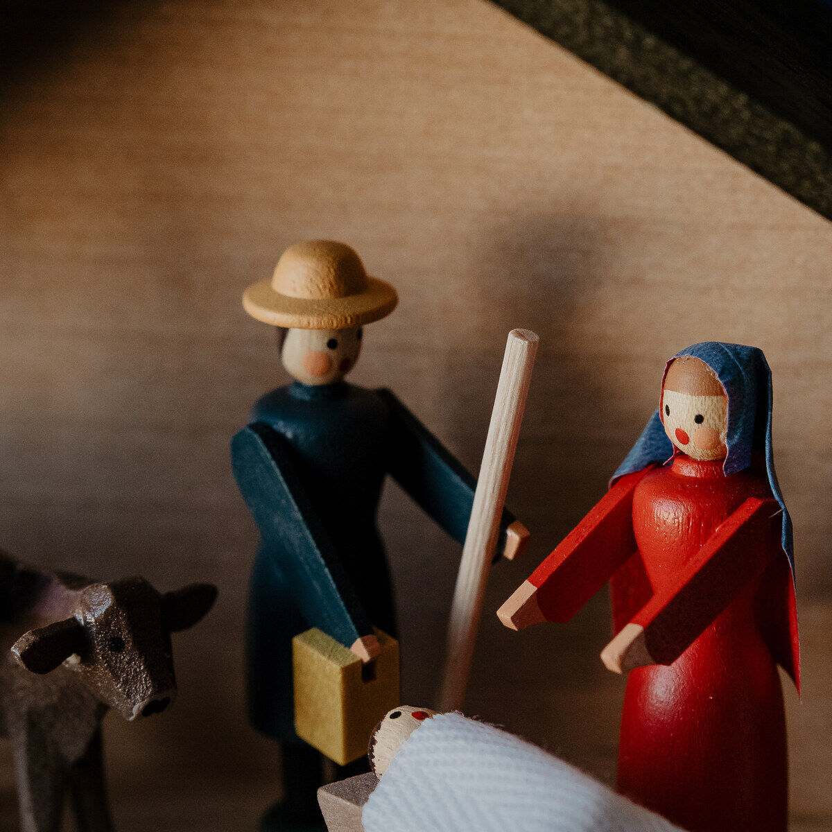 Detailaufnahme einer handgefertigten Weihnachtskrippe aus Holz. Zu sehen sind Figuren der Heiligen Familie, ein Esel und ein Stier in einem hölzernen Stall. Maria trägt ein rotes Gewand mit blauem Kopftuch, Josef ein dunkles Gewand, und das Christkind liegt in der Krippe. Eine besinnliche Darstellungsweise der Geburt Jesu, perfekt für die Weihnachtsdekoration.