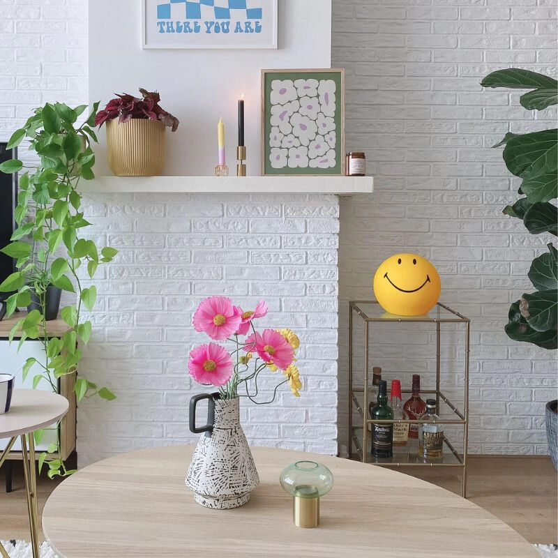 Wohnzimmer mit moderner Dekoration: Auf einem goldenen Beistelltisch steht eine leuchtende, gelbe LED-Lampe in Smiley-Form, daneben mehrere Flaschen. Im Vordergrund befindet sich ein runder Holztisch mit Vase und rosa Blumen. Im Hintergrund Pflanzen, weiße gemauerte Wand, Bilderrahmen, Kerzen und weitere Dekoartikel.