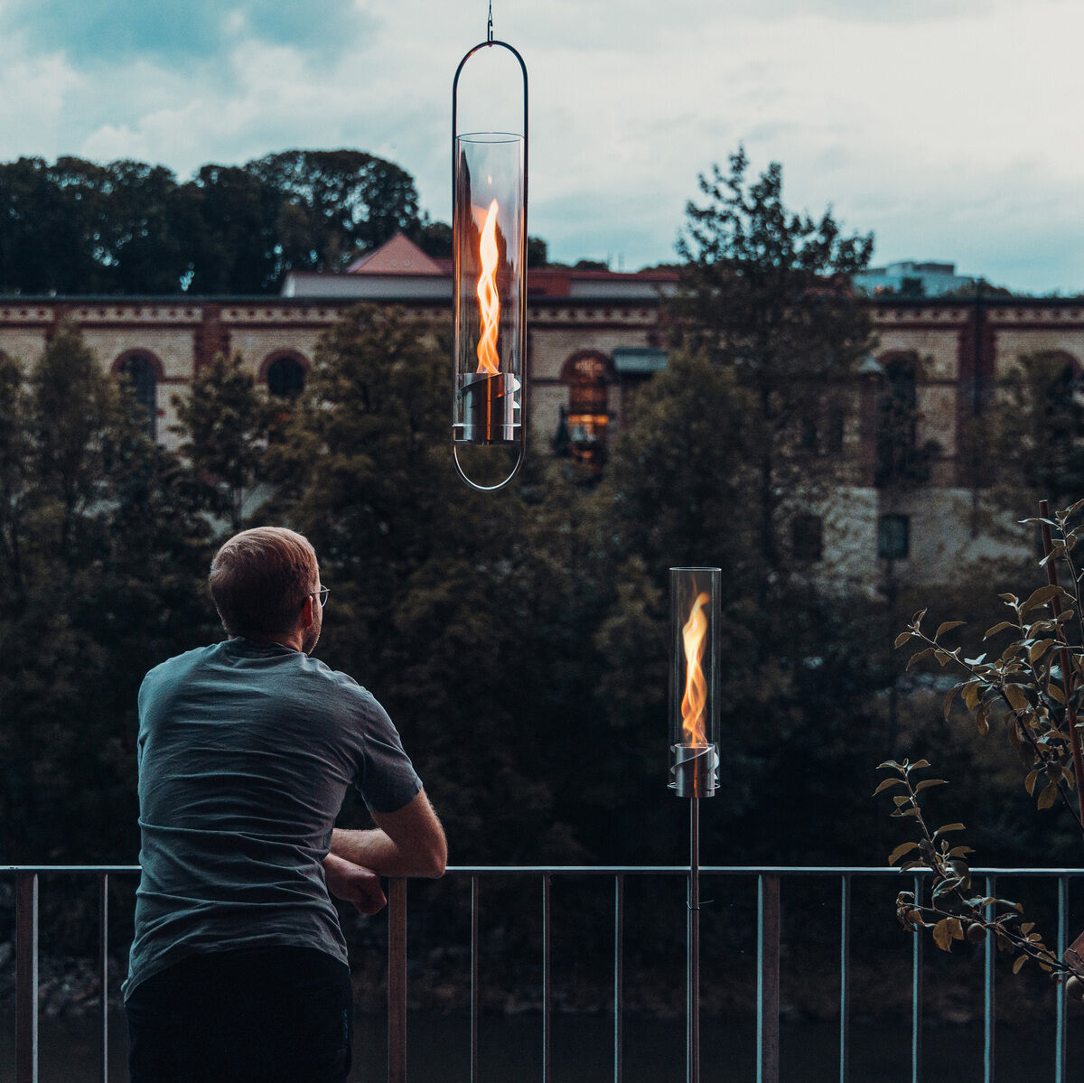 Ein Mann steht auf einer Terrasse und schaut auf eine moderne Gartenbeleuchtung. Zwei SPIN Erdspieße mit Flammenlicht leuchten im Vordergrund, und im Hintergrund ist eine städtische Landschaft mit Gebäuden und Bäumen zu sehen. Der Himmel ist bewölkt und das Ambiente ist stimmungsvoll und entspannend.
