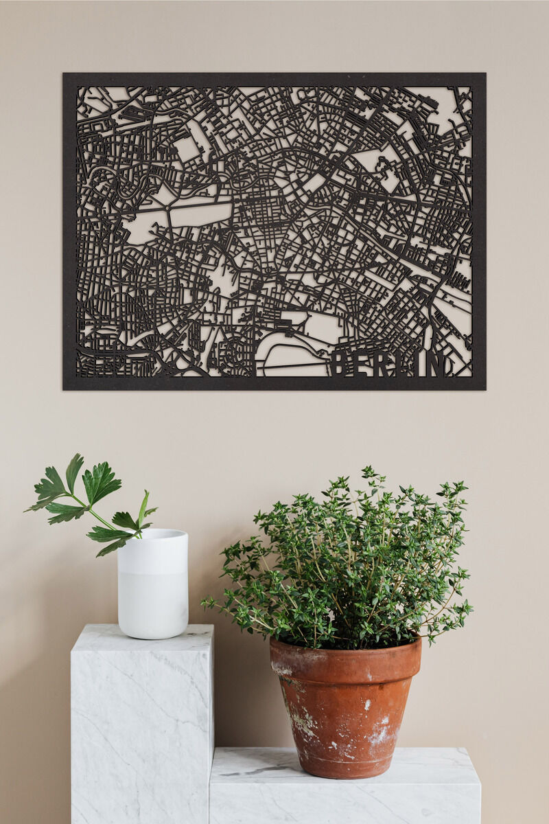 Ein kunstvoller, detaillierter Stadtplan von Berlin aus Holz, in Schwarz gehalten, hängt an einer beigen Wand. Die Karte wird von zwei Pflanzen eingerahmt, eine in einem weißen Keramiktopf und eine in einem Terrakottatopf.