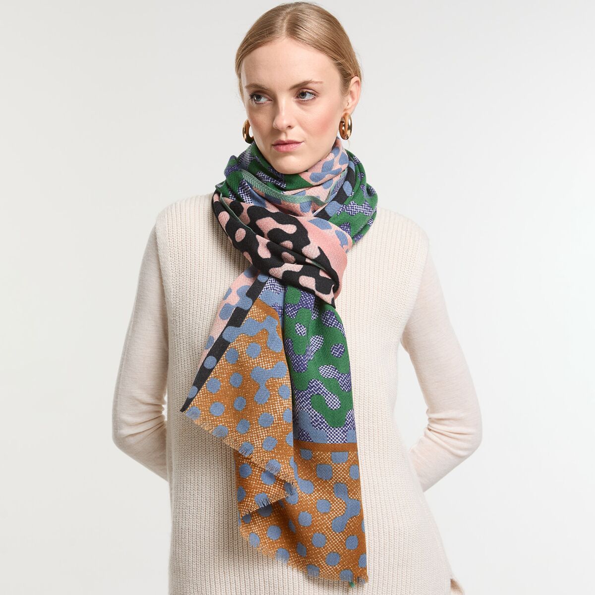 Eine Frau trägt einen langen, gemusterten Wollschal in Grün- und Brauntönen mit geometrischen und gepunkteten Mustern. Der Schal ist modisch und wirkt warm. Sie trägt dazu einen hellen, schlichten Pullover und steht vor einem neutralen Hintergrund.