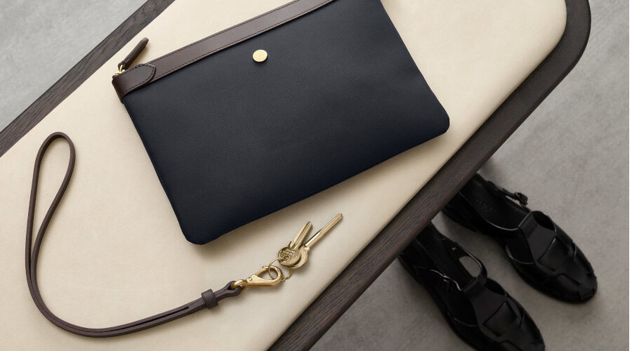 Schwarze Leder-Clutch mit braunem Reißverschluss und Schlüsselanhänger auf einem hellen Stuhl, daneben schwarze Sandalen