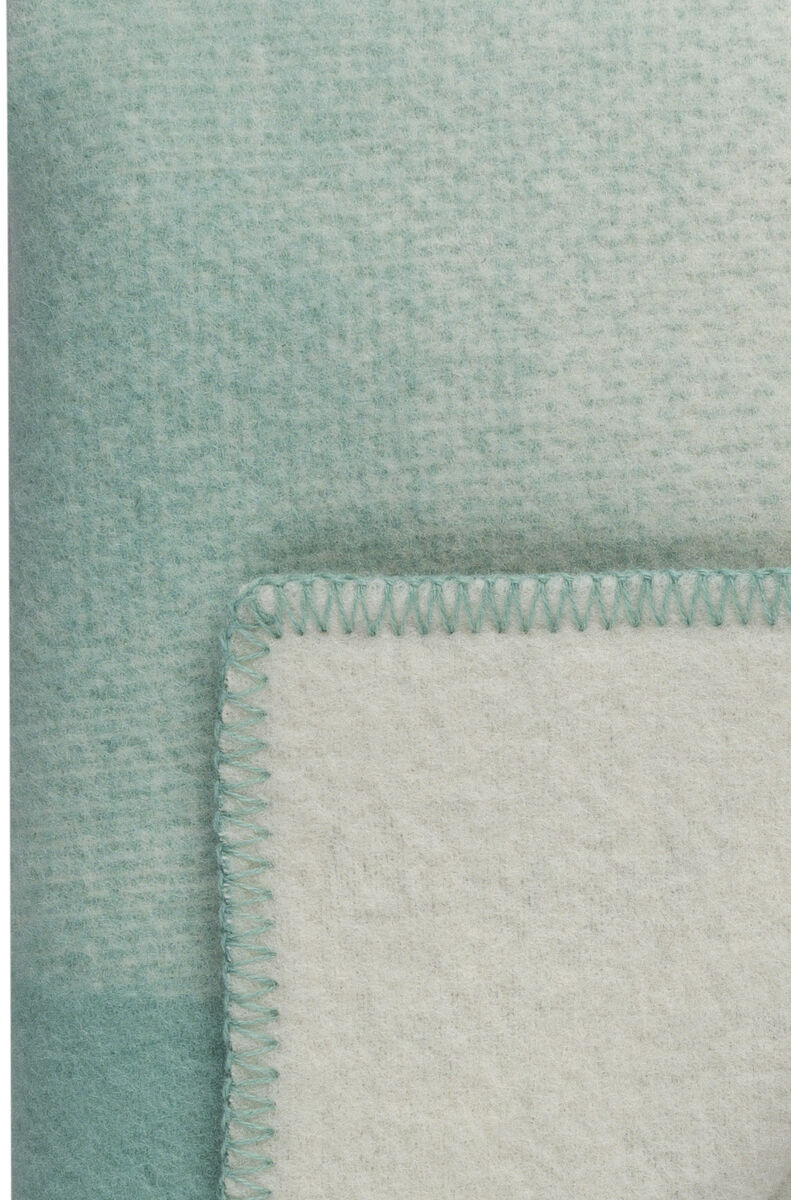 Eine hochwertige Lammwolldecke in hellgrüner und weißer Farbe ist gefaltet und auf einer weißen Fläche platziert.