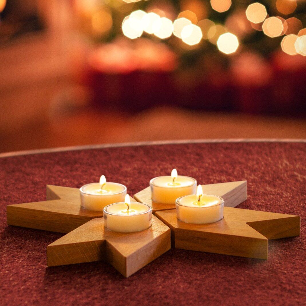 Adventskranz in Sternform mit vier brennenden Teelichtern aus Holz auf einem roten Tisch vor einem festlich beleuchteten Hintergrund.