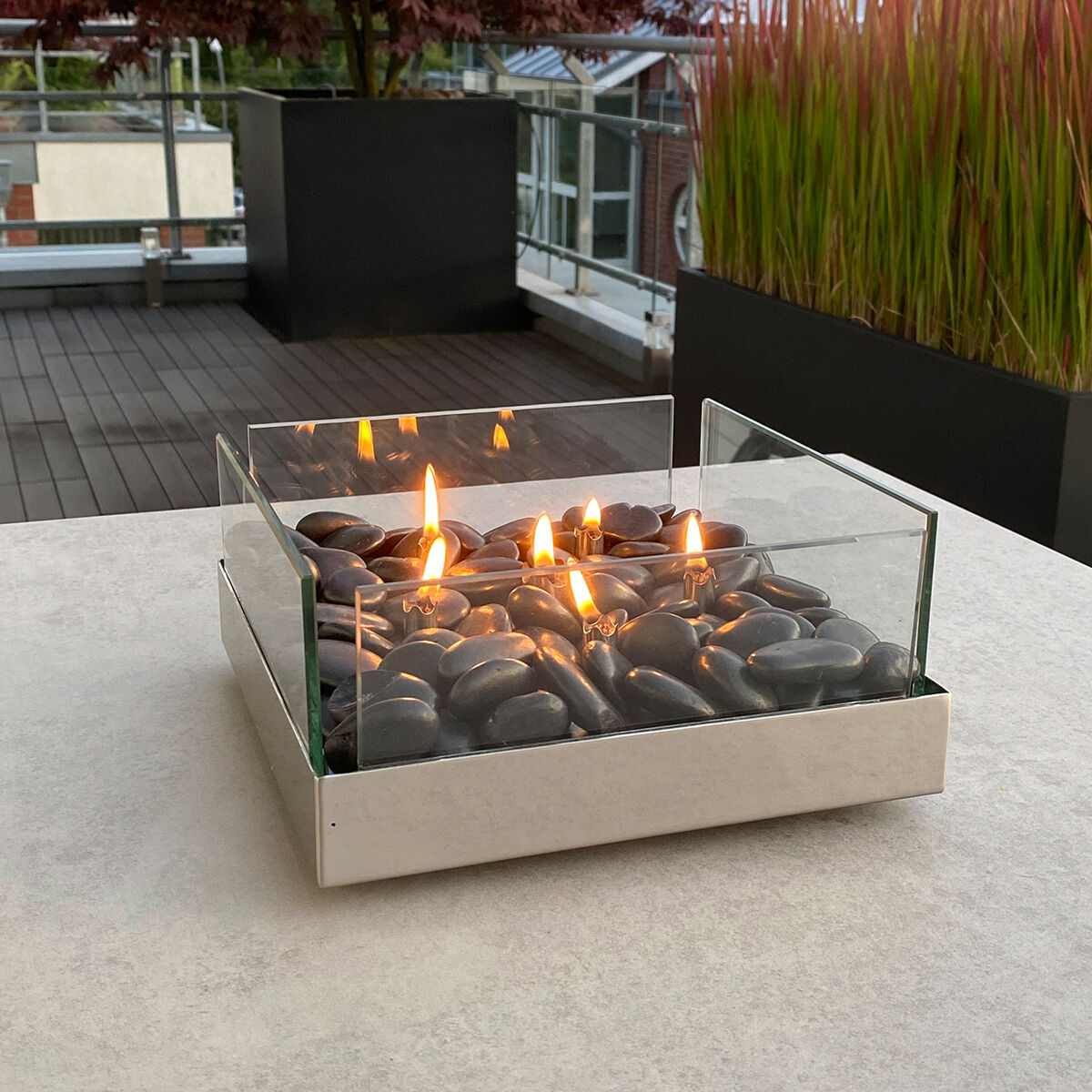 Ein Tischkamin mit Glasgehäuse und schwarzen Steinen auf einer Terrasse mit moderner Dekoration und brennender Flamme.