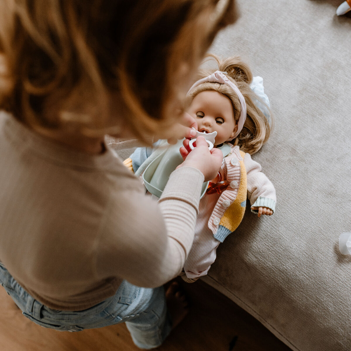Ein Kind spielt mit einer Badepuppe namens »Cosy Aquini Farbspiel«. Die Puppe liegt auf einem Sofa und wird mit einer kleinen Flasche gefüttert. Weitere Puppen und Zubehör sind in der Nähe zu sehen. Die Szene spielt in einem hell beleuchteten Kinderzimmer mit warmen Farben.