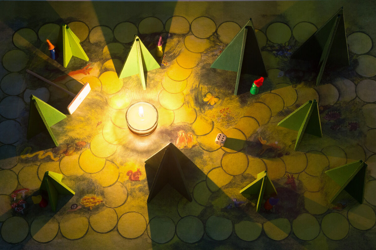 Ein Brettspiel namens 'Waldschattenspiel' zeigt ein beleuchtetes Spielbrett mit grünem Hintergrund, Kerze in der Mitte, kleinen Figuren und pyramidenförmigen Papierbäumen. Das Licht der Kerze erzeugt interessante Schattenmuster auf dem Spielbrett.