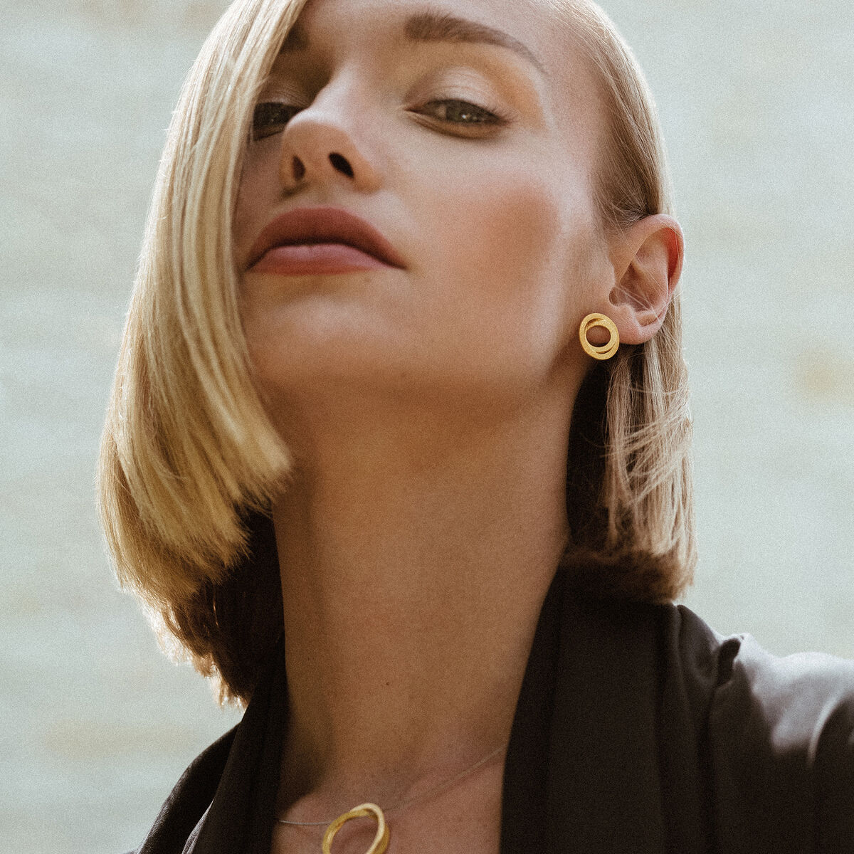 Eine Frau mit blonden Haaren trägt elegante silberne Ohrringe in Ringform. Sie trägt auch eine passende Halskette. Der Hintergrund ist unscharf und lichtdurchflutet, wodurch die Aufmerksamkeit auf den Schmuck gelenkt wird.