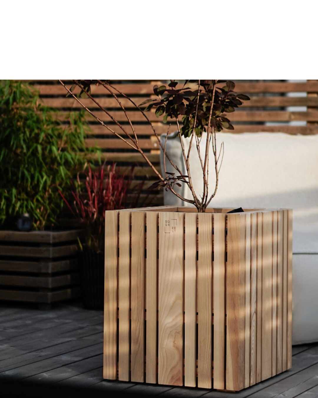 Ein Pflanzgefäß aus Holz, genannt 'GrowBIG', auf einer Terrasse. Das Pflanzgefäß hat vertikale Lamellen und enthält eine Pflanze mit roten Blättern. Im Hintergrund befinden sich weitere Pflanzen und ein weißes Polstermöbel.