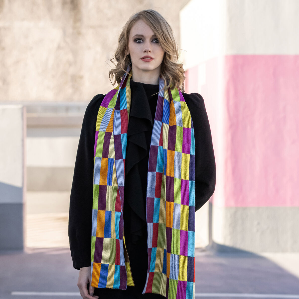 Eine junge Frau trägt einen langen Seidenschal von Ulrike Isensee Lager mit bunten, rechteckigen Mustern in Gelb, Rot, Lila, Türkis, Orange und Grün über einem schwarzen Outfit. Im Hintergrund sind moderne, farbig abgesetzte Wände und Schatten zu sehen.