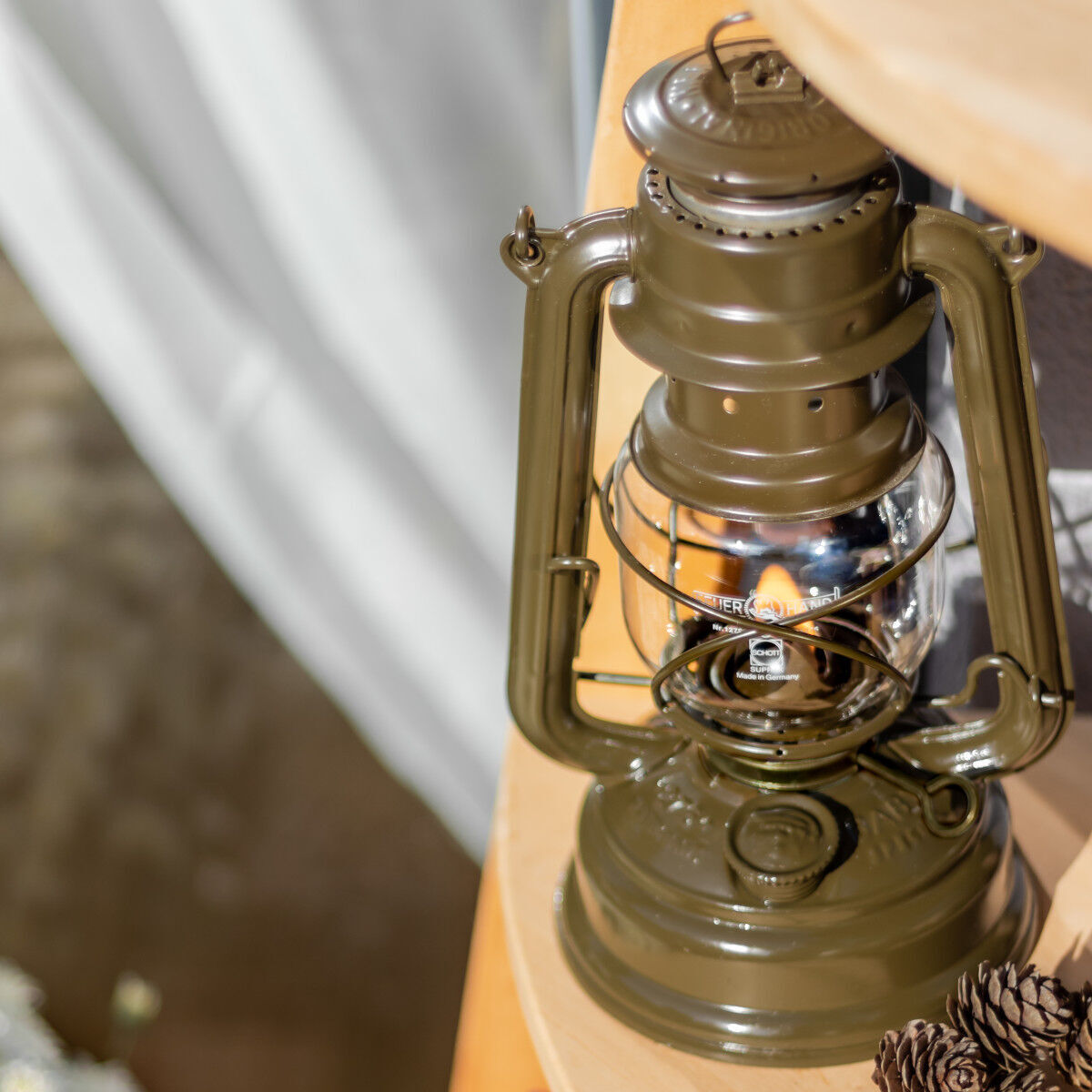 Eine stählerne Sturmlaterne auf einem runden, hölzernen Tisch. Die Laterne hat ein traditionelles Design und ist in den Farben Braun und Silber gehalten. Im Hintergrund sind unscharf Vorhänge zu erkennen.