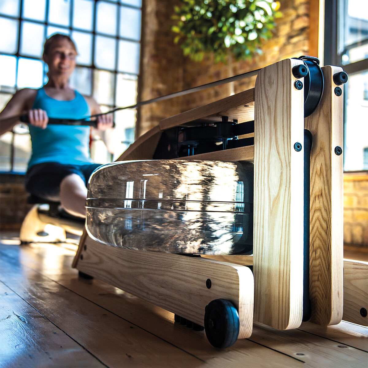 Ein elegantes Waterrower-Rudergerät aus Eiche in einem gut beleuchteten Raum. Im Hintergrund trainiert eine Person. Das Gerät ist aus naturbelassenem Holz gefertigt, mit einem transparenten Wassertank und Metallkomponenten, und steht auf einem Holzboden. Ein großes grünes Blattwerk ist ebenfalls sichtbar.