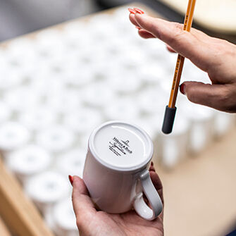 Hand hält weiße Tasse mit Text auf der Unterseite, andere Hand hält einen Stift über der Tasse