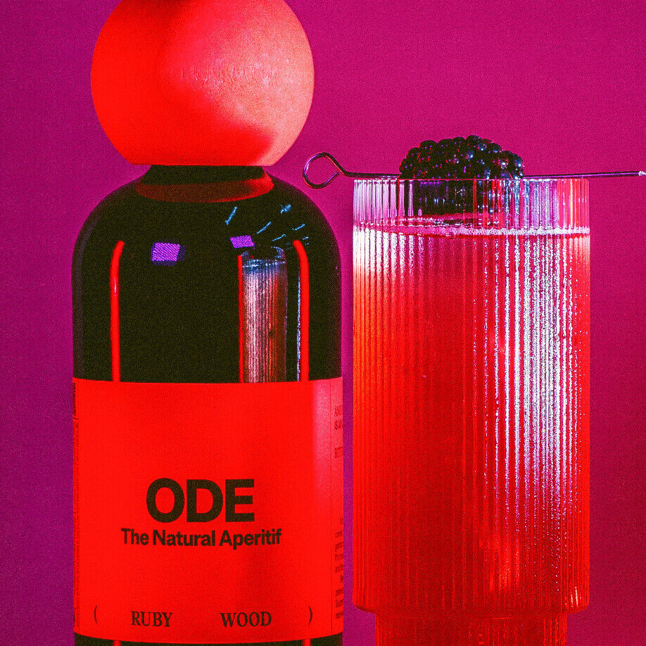 Bild einer eleganten Flasche mit roter Flüssigkeit und der Aufschrift 'ODE The Natural Aperitif Ruby Wood' neben einem Cocktailglas mit rotem Getränk und einer Brombeere auf einem Spieß. Hintergrund in kräftigem Rosa gehalten.