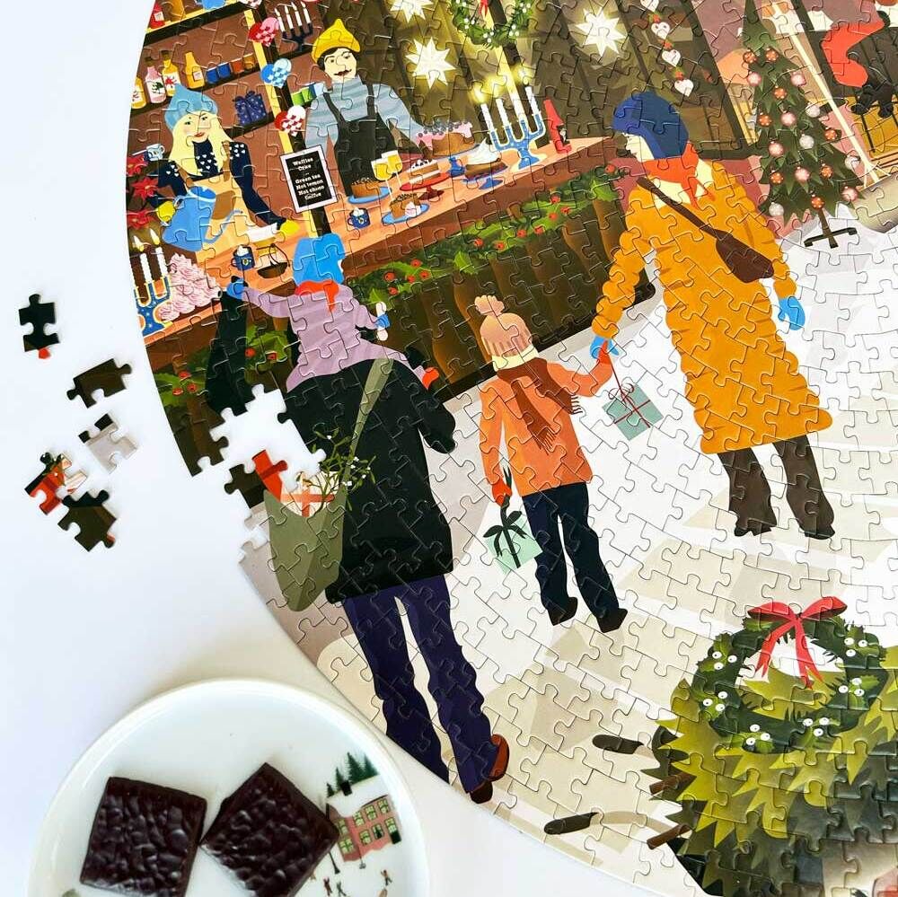 Das Bild zeigt ein zusammengesetztes Puzzle mit einem Weihnachtsmarkt-Motiv. Man sieht eine Familie bestehend aus zwei Erwachsenen und einem Kind, die einen festlich dekorierten Marktstand betrachten, umgeben von weihnachtlicher Beleuchtung und Dekorationen. Im Vordergrund des Bildes liegen Schokolade und eine weihnachtliche Zwergfigur.