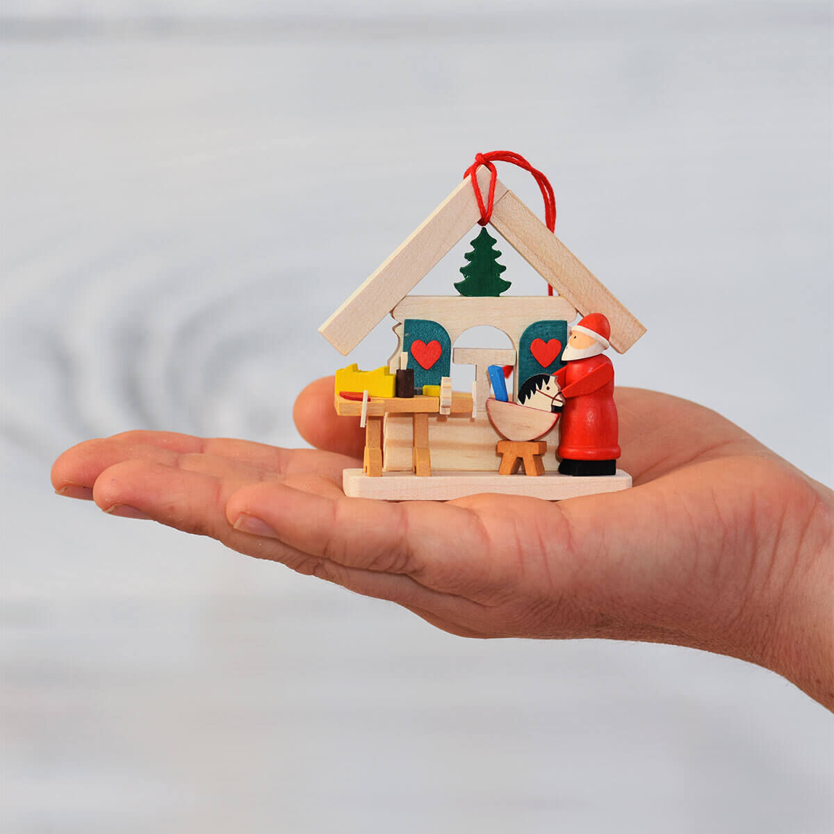 Ein Hand hält ein dreiteiliges Baumschmuck Geschenk-Set in Form von kleinen Weihnachtsmannhäusern. Die Dekorationen bestehen aus Holz und sind farbenfroh bemalt mit Weihnachtsmotiven, darunter rot, grün, weiß und Naturholz. Ein rotes Band dient zum Aufhängen.