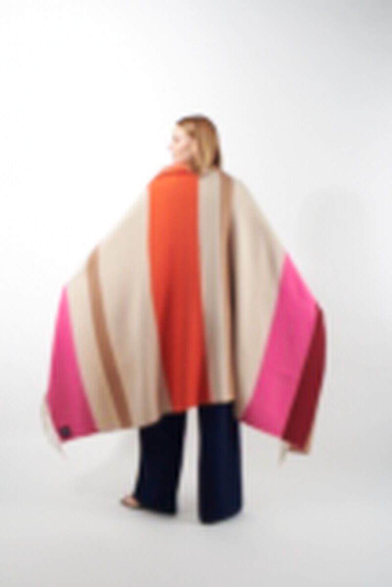 Eine Frau trägt ein Cape aus Alpaka-Wolle namens »Lucma« mit breiten Streifen in den Farben Beige, Orange, Braun, Pink und Rot. Sie steht vor einem weißen Hintergrund und das Cape breitet sich weit hinter ihrem Rücken aus.