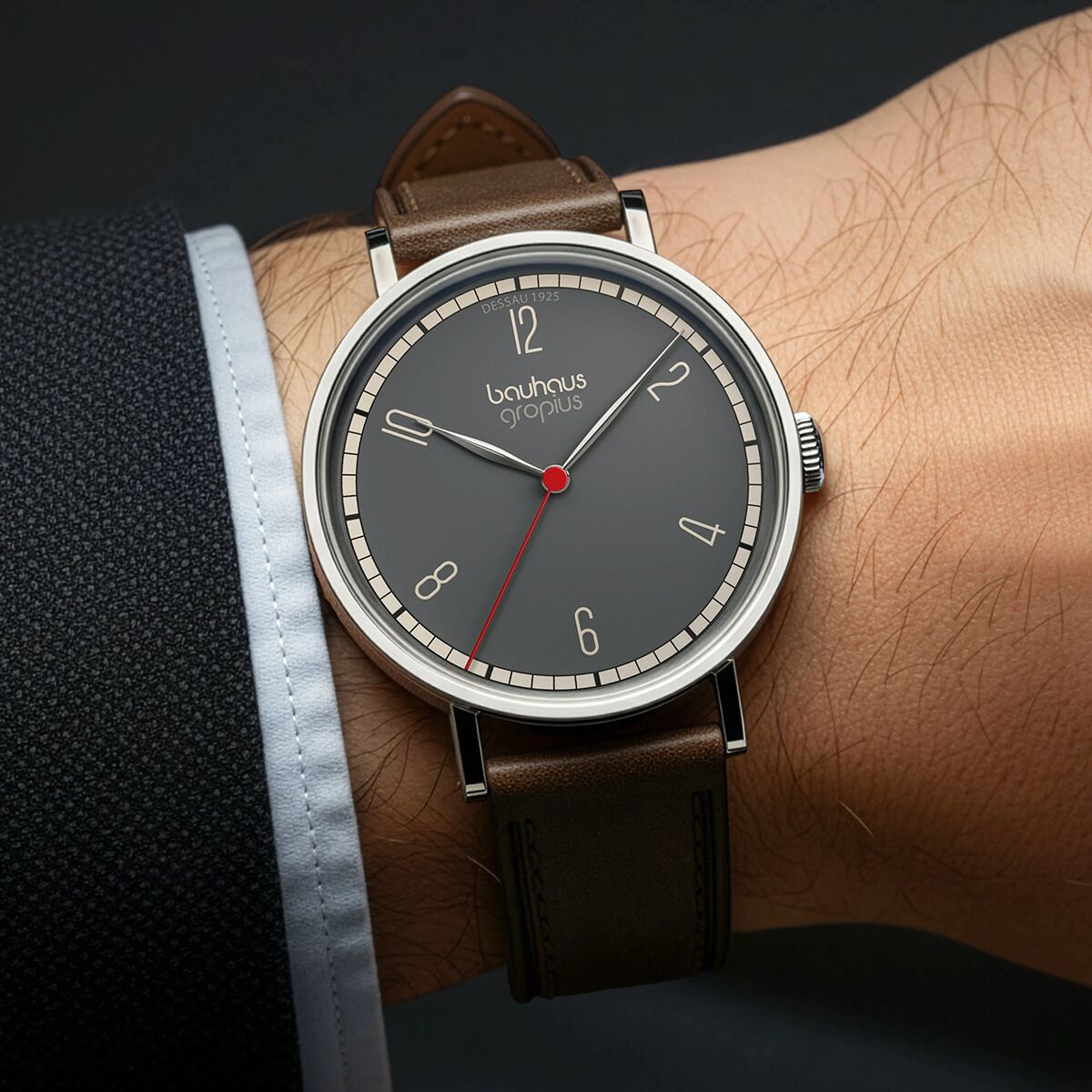 Armbanduhr im Bauhaus-Stil mit braunem Lederarmband, silbernem Gehäuse und grauem Zifferblatt, getragen am Handgelenk einer Person. Die Uhr zeigt ein minimalistisches Design mit roten, weißen und grauen Details.