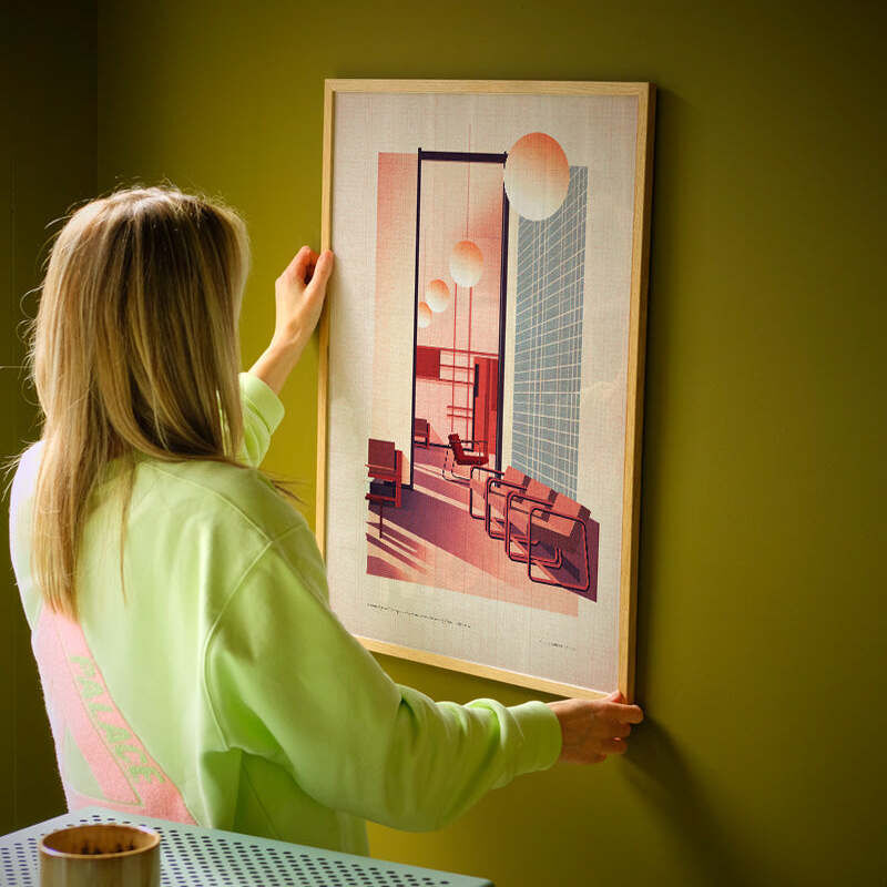 Eine Person hängt ein Poster mit einem modernen Architekturmotiv der Bocconi Universität Mailand an eine grüne Wand. Der Druck zeigt eine Innenansicht mit stilisierten Gebäudeteilen, geometrischen Linien und warmen Farbtönen. Im Vordergrund steht ein kleines Möbelstück mit einer Tasse darauf. Das Bild vermittelt Urbanität und modernes Design.
