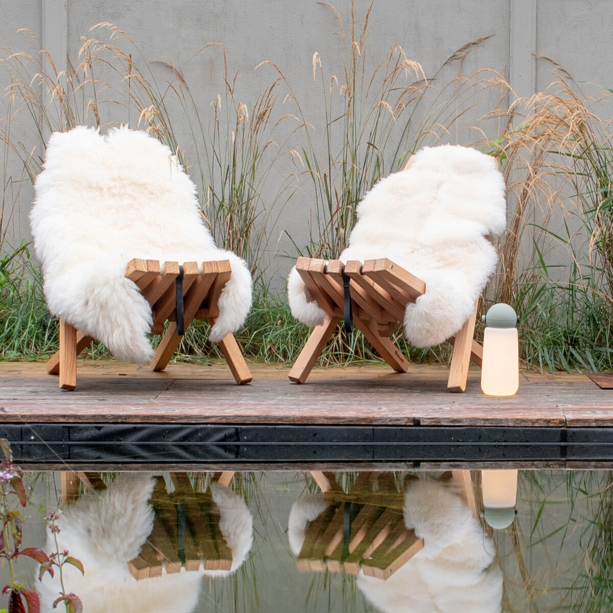 Zwei Holzstühle mit Schafsfellen bedeckt stehen nebeneinander auf einer Holzterrasse neben einem Ofen und einem Holzregal. Im Hintergrund Gräser und eine Wasserfläche.