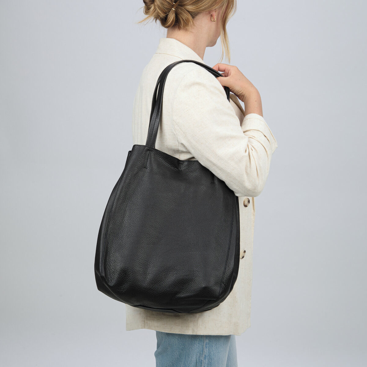 Elegante schwarze Lederhandtasche der Marke Guerer & Guerer mit strukturierter Oberfläche, getragen von einer Frau im cremefarbenen Mantel und Jeans. Die Tasche besitzt zwei lange Henkel und präsentiert sich schlicht sowie hochwertig verarbeitet. Hintergrund einfarbig hell.