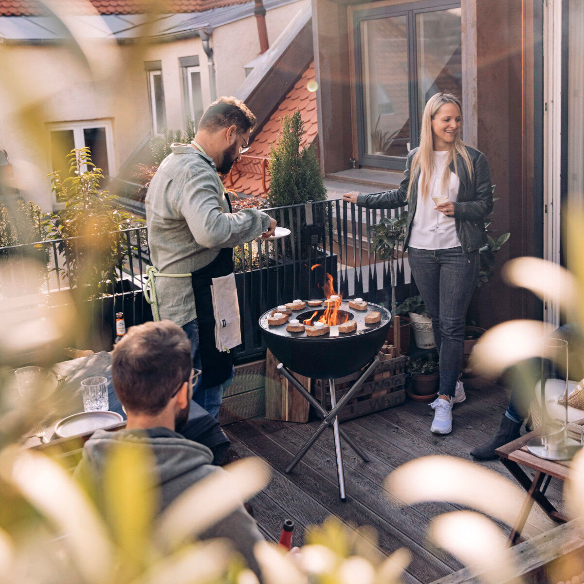 Personen genießen eine Grillparty auf einer Terrasse mit einem BOWL Plancha Halbring. Die Szene zeigt eine gemütliche Atmosphäre mit Freunden und Essen, das auf dem Grill zubereitet wird. Pflanzen und architektonische Elemente des Hauses sind im Hintergrund sichtbar.