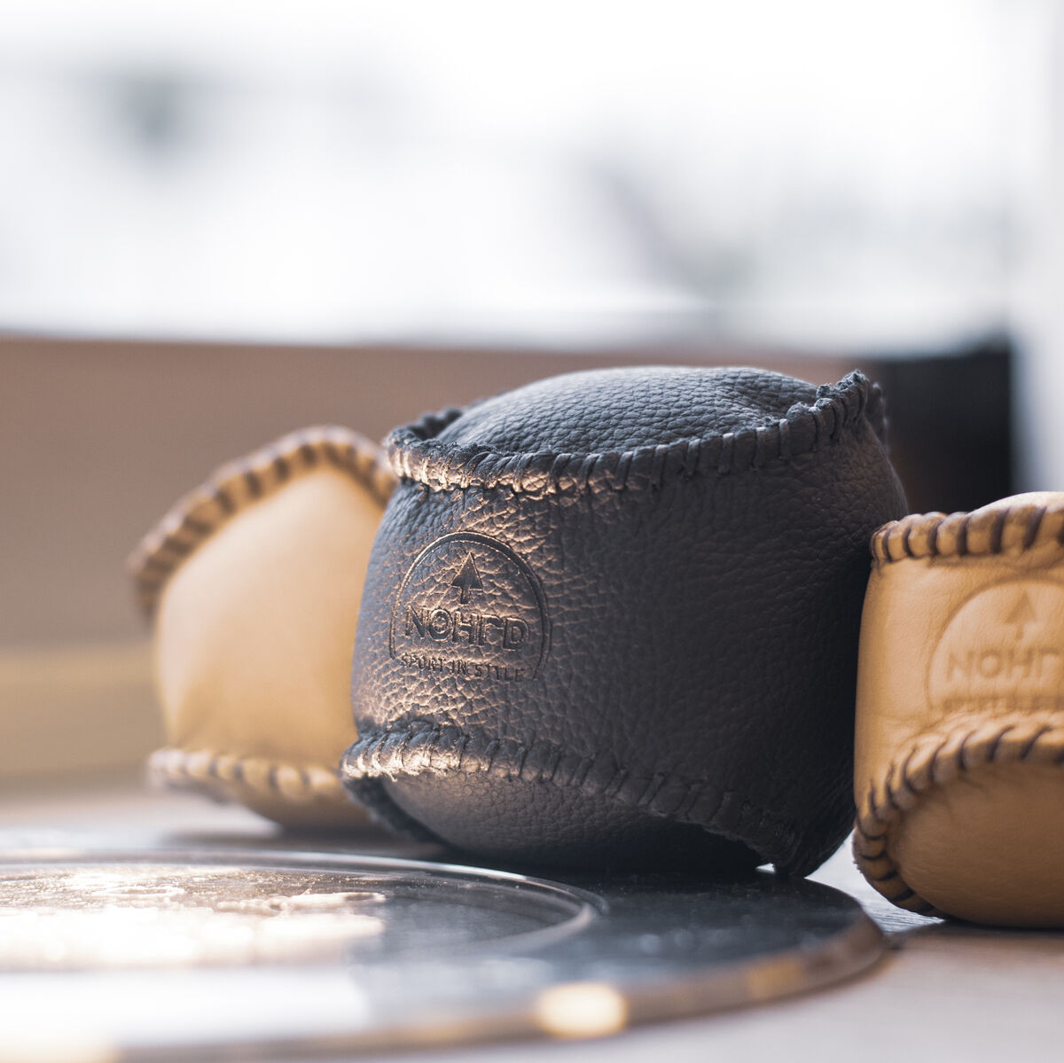 Ein HaptikBall in Schwarz, gefertigt aus Leder, steht im Vordergrund, flankiert von zwei weiteren Bällen aus beigefarbenem Leder. Sie liegen auf einem runden Untersetzer auf einem Holztisch, was eine gemütliche, natürliche Atmosphäre erzeugt.