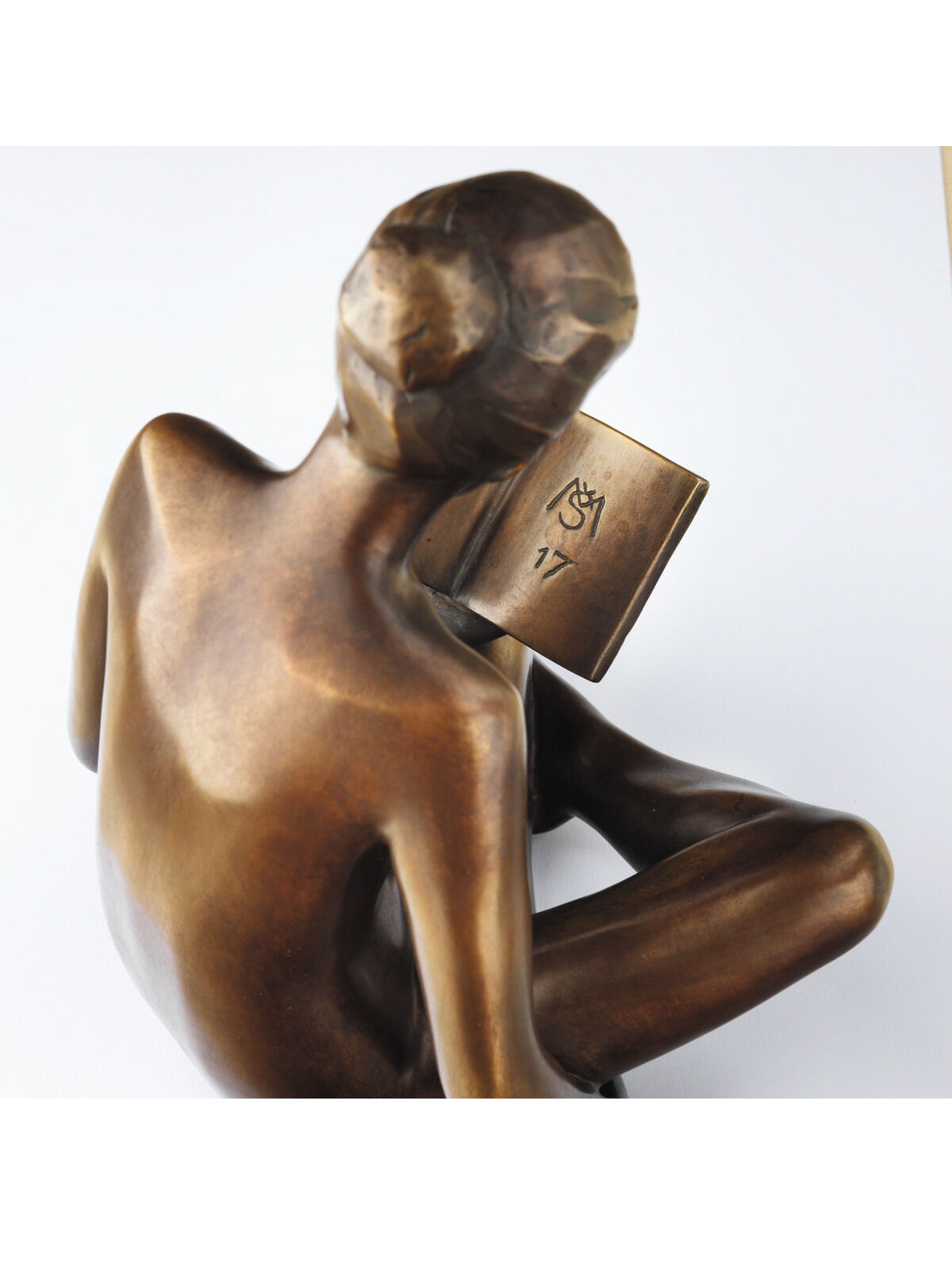 Bronzeskulptur einer sitzenden Person, die ein Buch liest. Die Oberfläche hat eine glatte, polierte Erscheinung mit natürlichen Brauntönen. Die Figur ist in einer geschlossenen, nachdenklichen Pose gehalten.