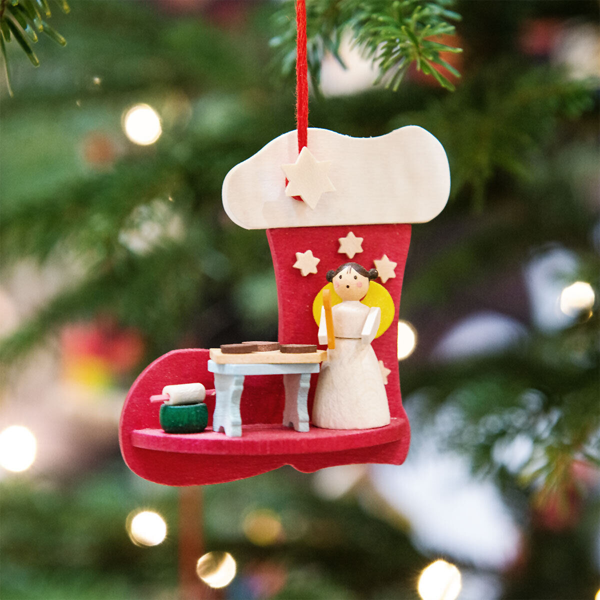 Baumschmuck in Form eines roten Strumpfes mit einem handgefertigten Engel, der auf einem Podest steht, und weiteren weihnachtlichen Details, hängend an einem Weihnachtsbaum.
