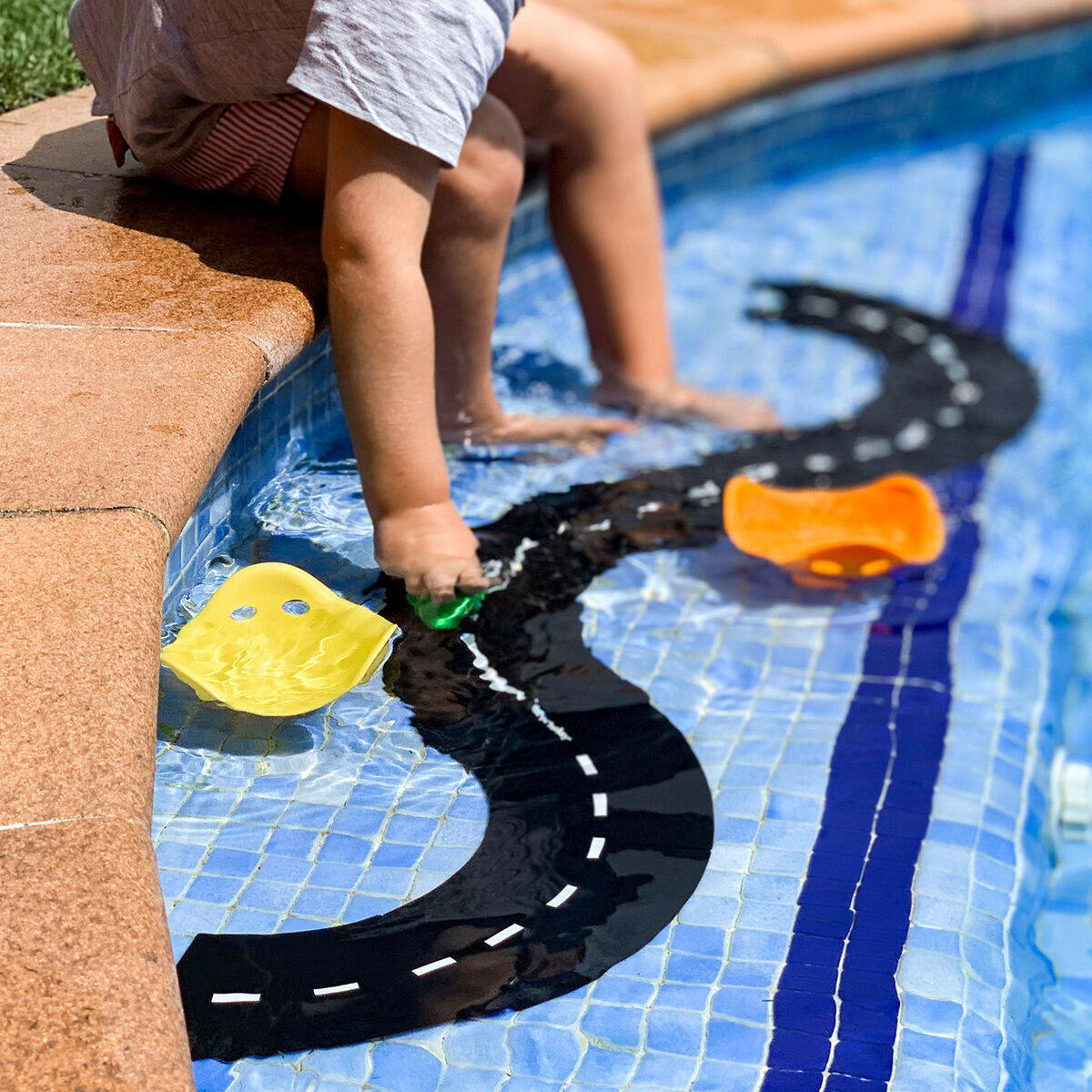 Ein Kind sitzt am Rand eines Pools und spielt mit einem Straßenbau-Set »Ringroad«. Schwarze, flexible Straßenteile mit weißen Markierungen schwimmen auf dem Wasser, während bunte Boote daneben schwimmen. Im Hintergrund ist ein Handtuch auf grünem Rasen sichtbar. Die Szene findet bei Tageslicht im Freien statt und vermittelt sommerliche Stimmung und Spielspaß am Wasser.