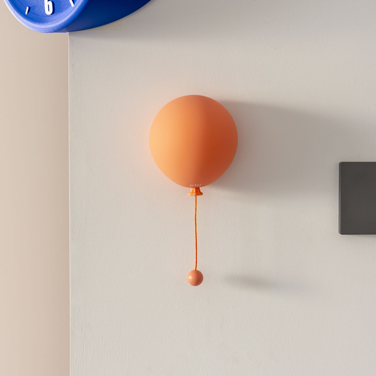 Eine orangefarbene Wandleuchte »Mimo Ballon« von Gingko Design Ltd in Ballonform hängt an einer cremeweißen Wand. Darüber ist eine blaue Wanduhr zu sehen. Die Szene wirkt modern und hell, ideal für stilvolle Wohn- oder Kinderzimmer.