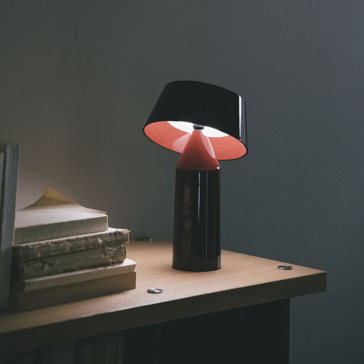Eine moderne Leseleuchte »Bicoca« steht auf einem Holztisch neben einem Stapel Bücher. Die Lampe hat einen schlichten und eleganten Stil mit einem schwarzen und roten Design. Weiches Licht leuchtet den dunklen Raum leicht aus.