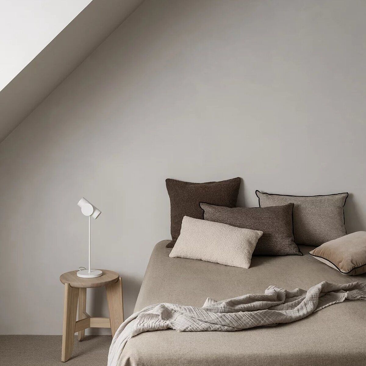 Helles, minimalistisch eingerichtetes Schlafzimmer mit Sitzhocker „ELI“ aus hellem Holz neben einem Bett mit neutralen Kissen und Decke. An der Wand befinden sich dekorative Holzbalken, das Tageslicht fällt durch ein großes Dachfenster. Das Ambiente ist ruhig, modern und naturverbunden.