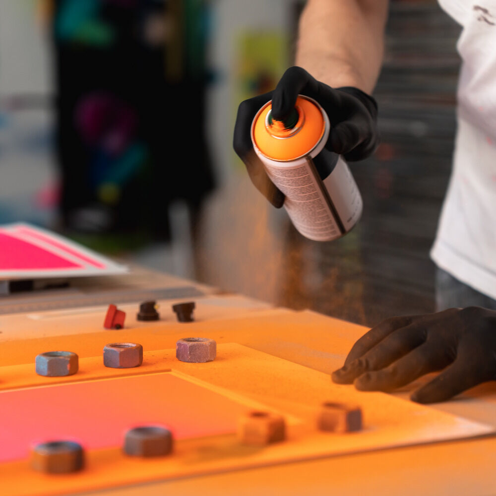 Ein Künstler mit schwarzen Handschuhen sprüht orangenen Lack auf ein kunstvoll vorbereitetes Siebdruckpapier. Farbtöpfe, Siebdruckrahmen und kräftige Farbverläufe in Blau, Orange und Pink sind auf einem Arbeitstisch angeordnet. Die Szene wirkt kreativ und experimentell.