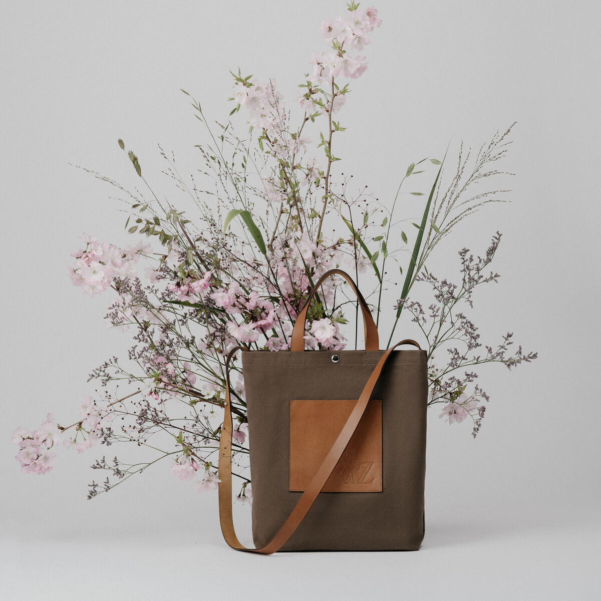 Ein minimalistischer ZEIT Shop Jubiläums-Shopper aus braunem Stoff, verziert mit einem Blumenarrangement aus rosa und grünen Blumen, vor einem grauen Hintergrund.