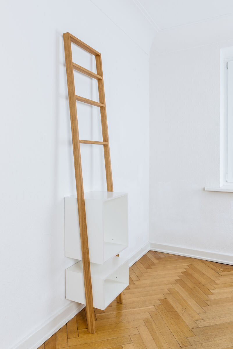 Ein Leiterregal mit der Bezeichnung »brecht«, das aus einer Holzleiterkonstruktion und weißen Regalboxen besteht, steht an einer Wand in einem Raum mit Fischgrätenparkett und weißen Wänden.