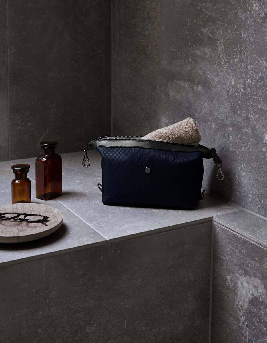 Eine hochwertige, schwarze Kulturtasche von Mismo auf einer grauen Steinplatte im Badezimmer. Neben der Tasche befinden sich zwei braune Glasfläschchen und eine aufgerollte Handtuch.