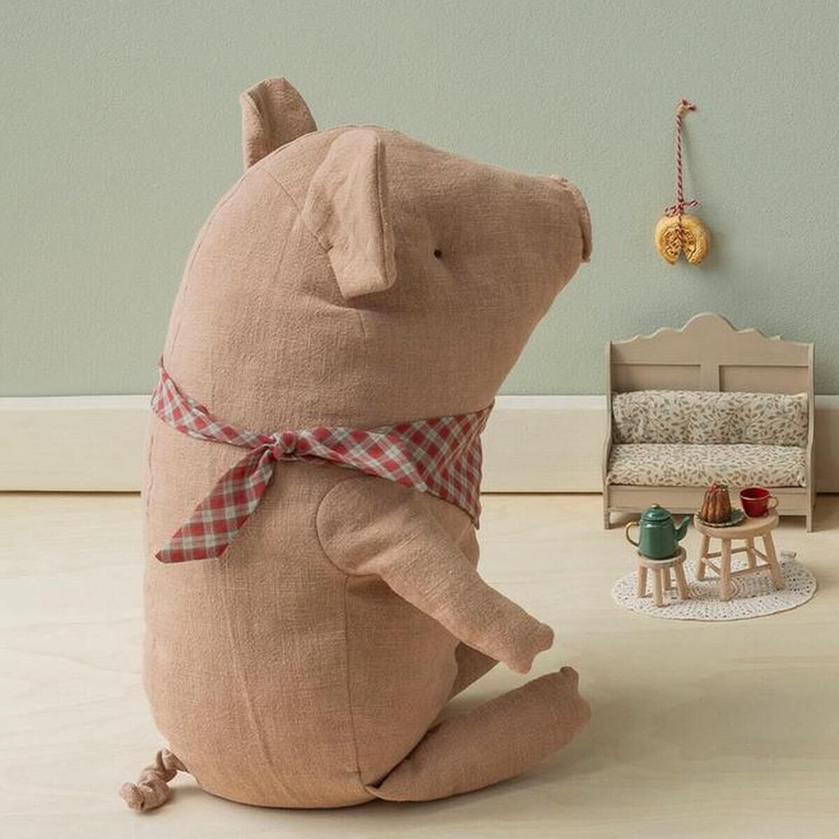 Das Bild zeigt das Maileg Kuscheltier »Großes Trüffelschwein« sitzend mit kariertem Schal in beige-rosafarbenem Stoff in einem kindlich dekorierten Raum. Im Hintergrund sind kleine Stofffiguren, Miniaturmöbel und Deko, alles in sanften Pastelltönen, arrangiert.