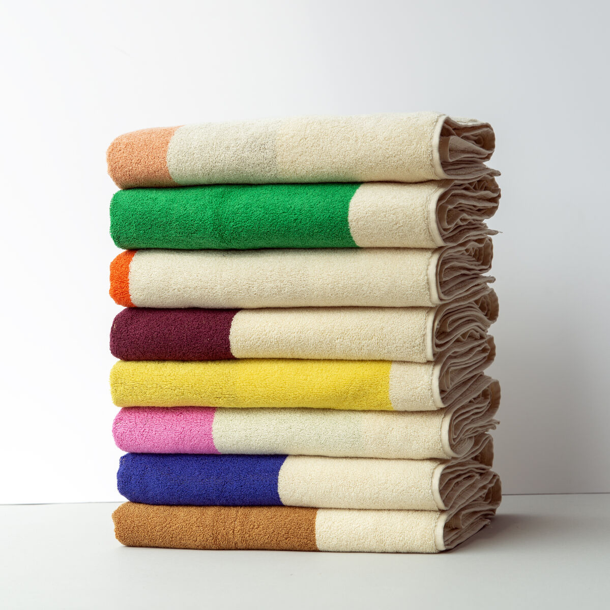 Ein Stapel von Handtüchern in verschiedenen Farben (Beige, Grün, Rot, Gelb, Lila, Blau, Orange), gefaltet und aufeinander gestapelt, vor einem weißen Hintergrund.