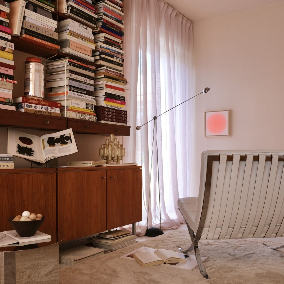 Helles, modern eingerichtetes Wohnzimmer mit Bücherregal, Designerstuhl, Vorhang in Pastellrosa und quadratischem Kunstdruck in Apricot an der Wand. Die Szene wirkt ruhig und harmonisch, geeignet als stilvolle Leseecke mit zeitgemäßer Wandgestaltung.