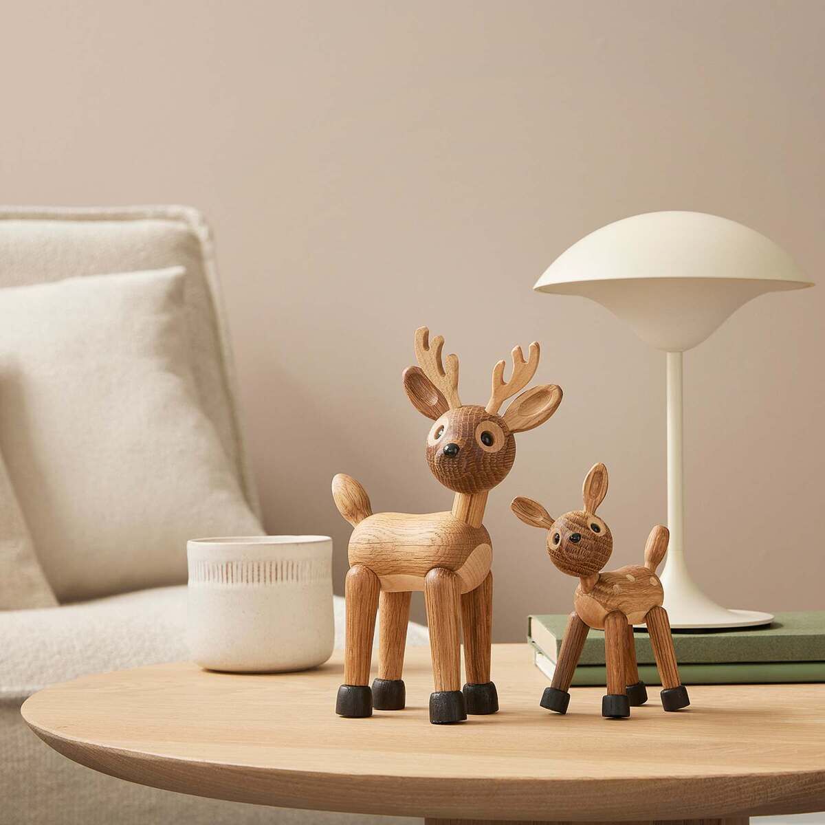 Zwei dekorative Holzfiguren in Rentierform stehen auf einem runden Holztisch in einem modern eingerichteten Wohnzimmer. Neben ihnen sind eine weiße Kerze, gestapelte Bücher und eine weiße Designlampe zu sehen. Im Hintergrund befindet sich ein helles Sofa vor einer beige gestrichenen Wand.