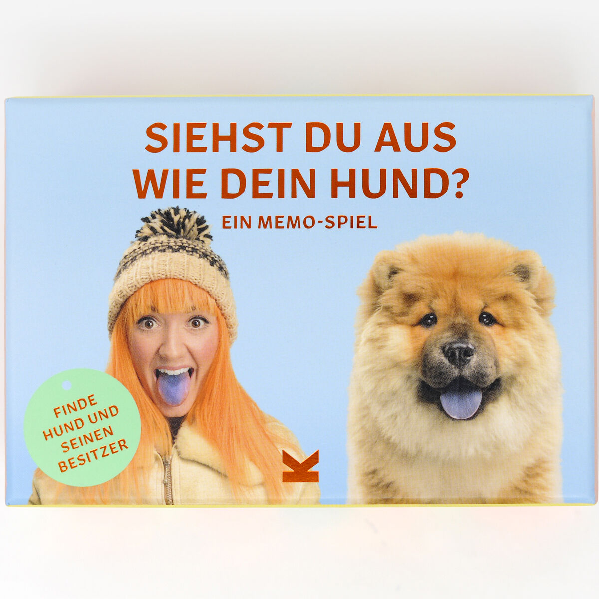Eine Spielschachtel mit dem Titel 'Siehst du aus wie dein Hund?' zeigt eine Frau mit orangen Haaren und einem Hund, die beide die Zunge herausstrecken. Das Spiel lädt dazu ein, Hunde ihren Besitzern zuzuordnen.