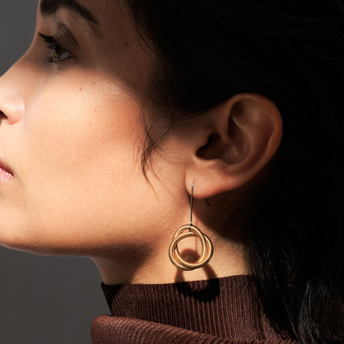 Nahaufnahme eines weiblichen Profils mit eleganten, vergoldeten Edelstahl-Ohrringen "Oh Tress Gold" von la molla. Die Ohrringe bestehen aus ineinandergreifenden, filigranen goldenen Coils und setzen dezente, architektonische Akzente. Sie werden zu einem braunen Oberteil getragen.