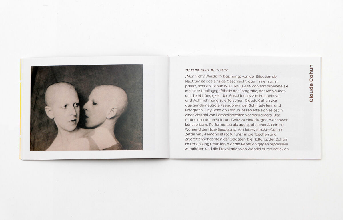 Dieses Bild zeigt das geöffnete Memo-Spiel „Fotografisches Gedächtnis“ auf einem weißen Hintergrund. Auf der linken Seite des Buchs ist eine schwarz-weiße Fotografie von zwei sich gegenüberstehenden Personen zu sehen. Die rechte Seite enthält Text über Claude Cahun.