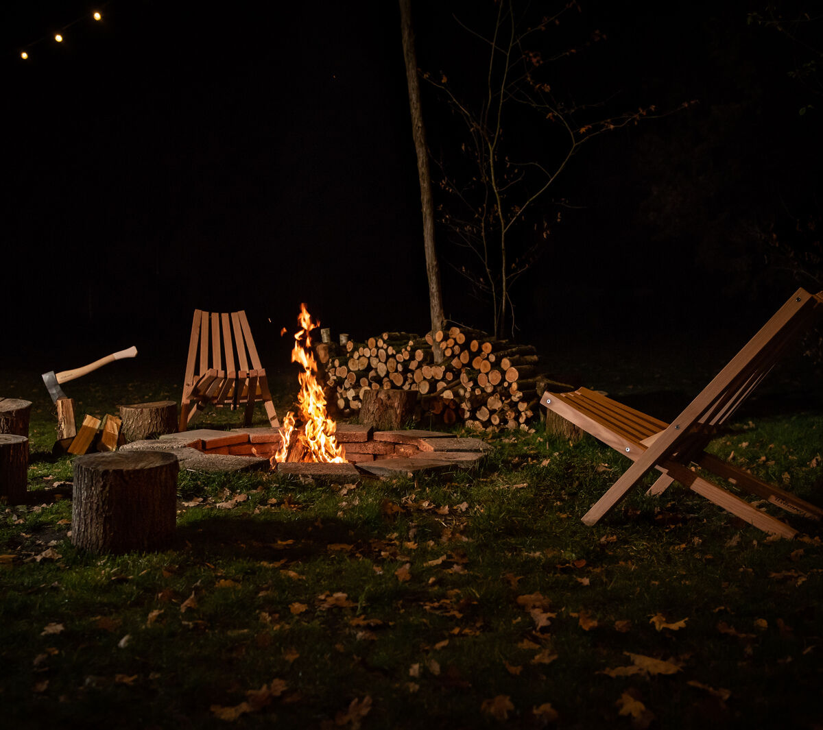 Ein stilvoller Feldstuhl mit Schafsfellauflage, geeignet für den Einsatz im Freien, steht neben anderen Holzstühlen und Baumstämmen um ein Lagerfeuer. Eine Axt liegt auf einem Baumstamm in einem natürlichen, ruhigen Gartenambiente unter Lichterketten.